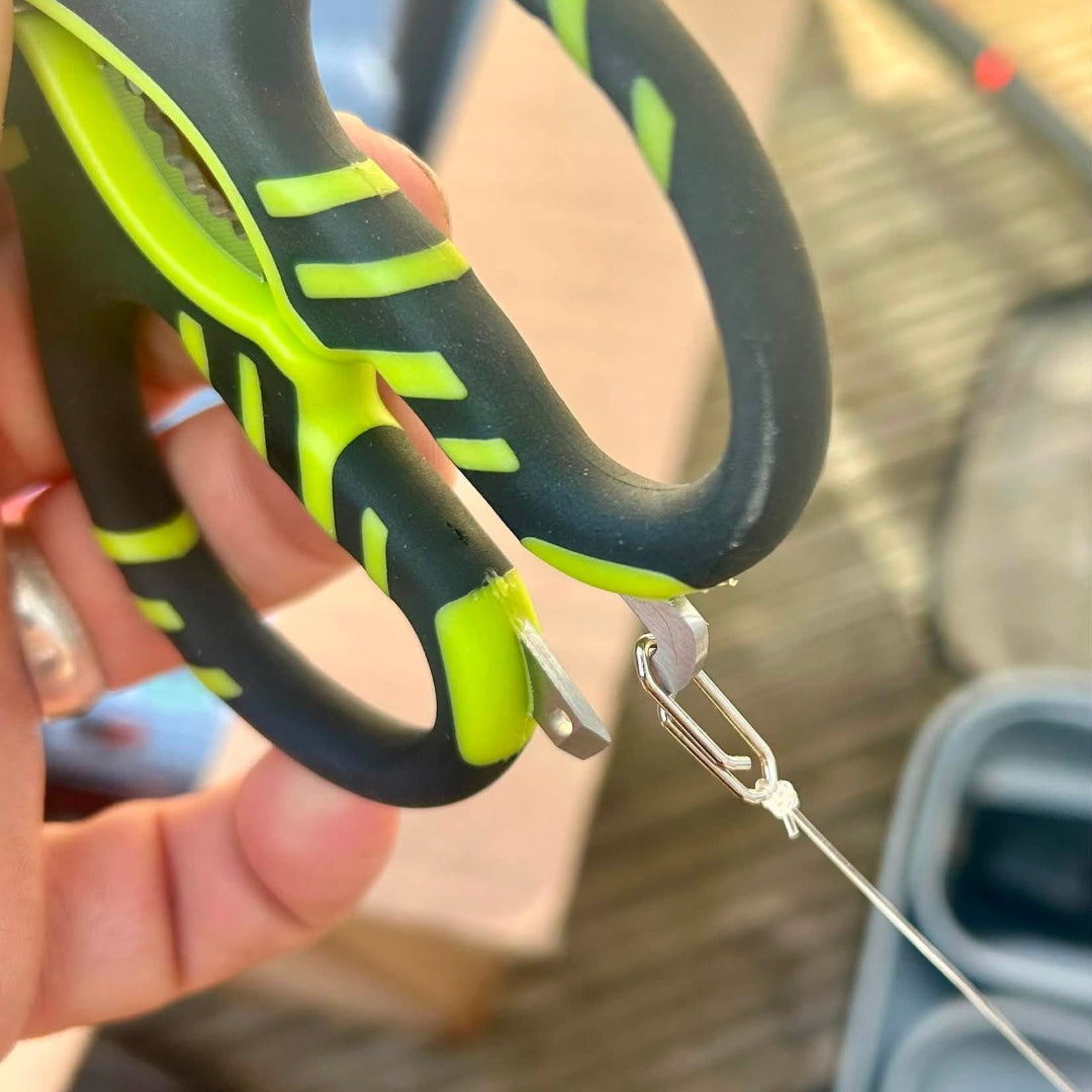 Close-up of a hand holding a pair of black and neon green utility scissors, using the metal blade to cut a fishing line attached to a carabiner.