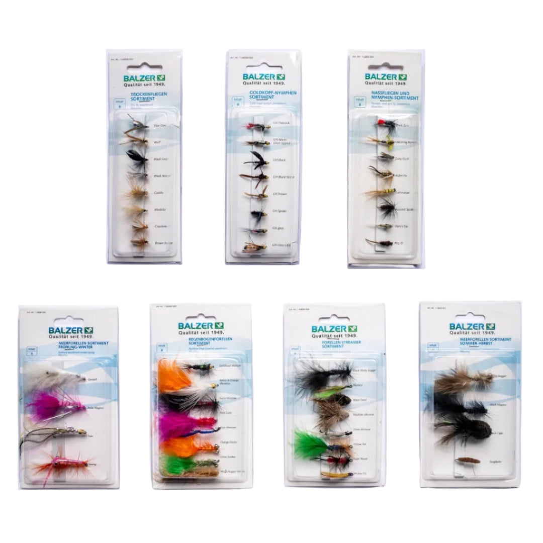 Seven packages of Balzer EJ fly fishing lures arranged on a white background, with three packages at the top and four at the bottom. The lures are shown through clear plastic blister packaging with printed labels and color-coded hooks.