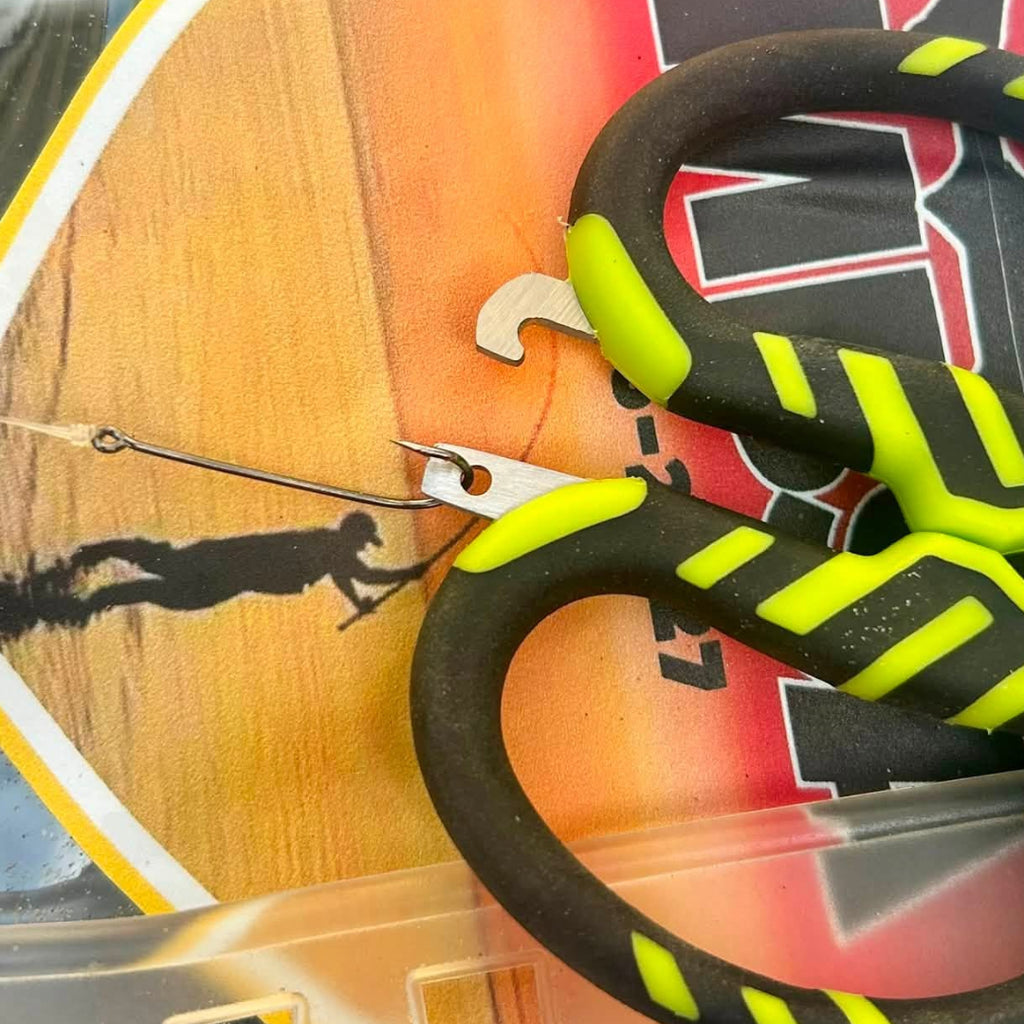 A close-up of a fishing hook set inside clear plastic packaging, featuring a bright green and black hook with a wire leader, placed over a red and yellow branded insert card. A black and neon yellow-handled tool is also visible in the foreground.