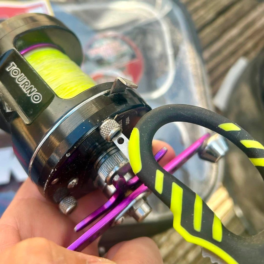 A close-up of a black spinning fishing reel with bright yellow-green line, held in a hand. The reel has a black metal body with silver knobs and a black handle featuring neon yellow-green accents.